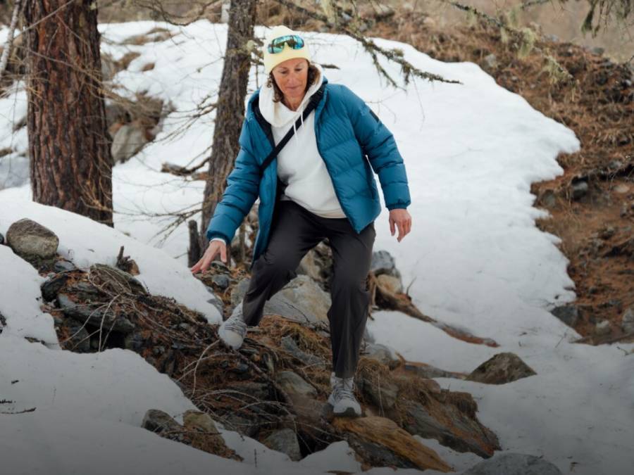 Two sleds with two people each are riding down a snowy slope. The people are laughing and wearing winter clothing, hats, and sunglasses; snow-covered mountains can be seen in the background.